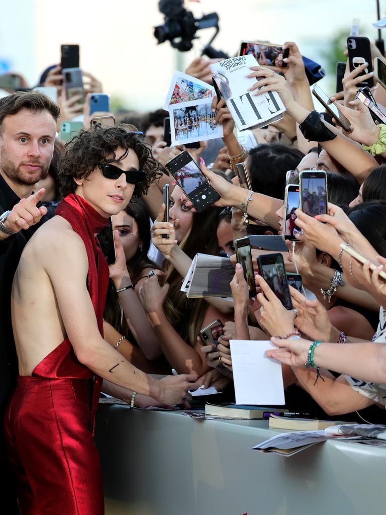 Timothée Chalamet steht vor einer Absperrung und signiert Autogramme, während auf der anderen Seite des Zauns dutzende Fans begeistert ihre Smartphones auf ihn richten.