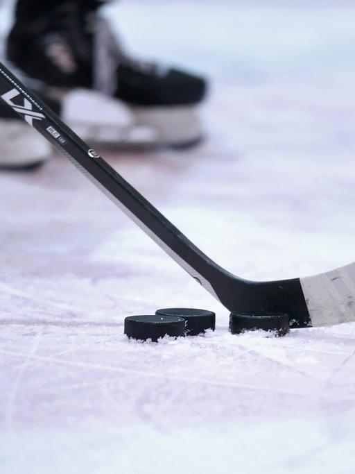 Ein Eishockeyschläger vor einem Puk auf dem Eis. Dahinter verschwommen ein paar Schlittschuhe.