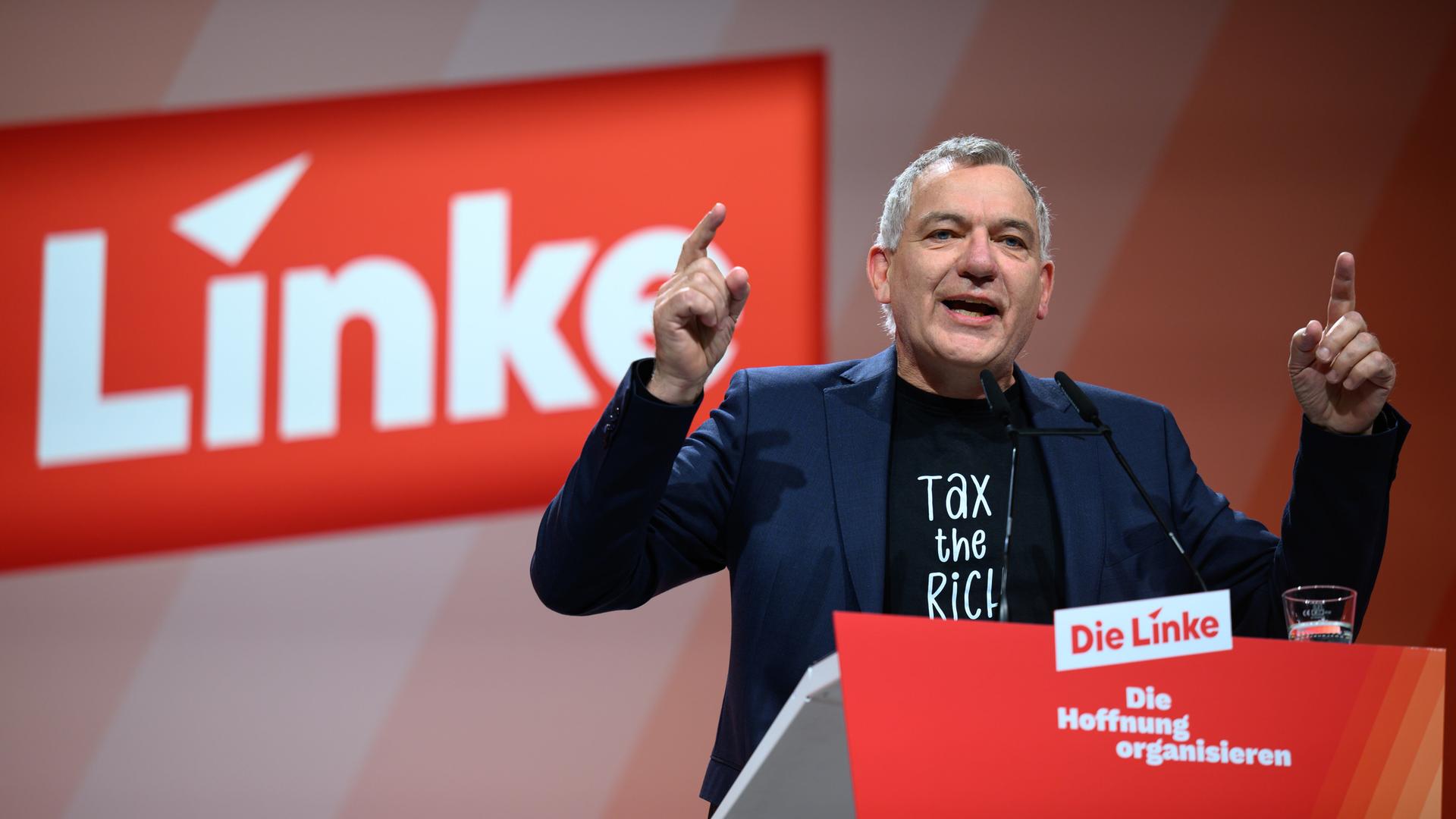 Jan van Aken, Parteivorsitzender Die Linke, spricht auf dem Bundesparteitag in Chemnitz. Er steht auf der Bühne und spricht in ein Mikrofon. Er trägt ein dunkles T-Shirt und ein blaues Sakko.