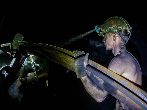 Ein mit Kohlenstaub bedeckter Bergmann trägt mit freiem Oberkörper ein schweres Metallrohr. Von seinem gelben Helm strahlt die Grubenlampe in die Dunkelheit der Kohlenmine. Stonava near Karvinâa, Czech Republicâ, January 22, 2025. 