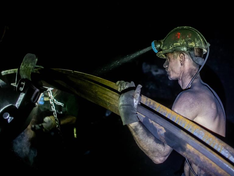 Ein mit Kohlenstaub bedeckter Bergmann trägt mit freiem Oberkörper ein schweres Metallrohr. Von seinem gelben Helm strahlt die Grubenlampe in die Dunkelheit der Kohlenmine. Stonava near Karvinâa, Czech Republicâ, January 22, 2025. 