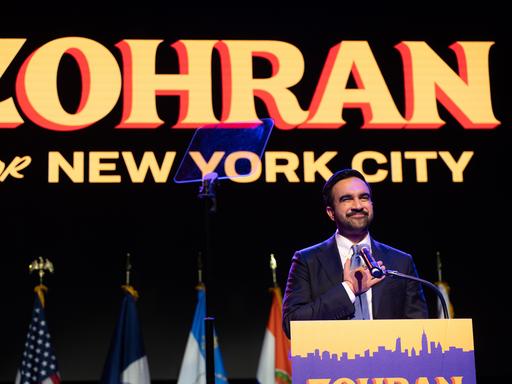 Ein Mann steht auf einer Bühne und fasst sich mit der rechten Hand aufs Herz. Es ist der neu gewählte New Yorker Bürgermeister Zohran Mamdani. Er hat einen Bart, hinter ihm ist ein Plakat mit seinem Vornamen aufgehängt.