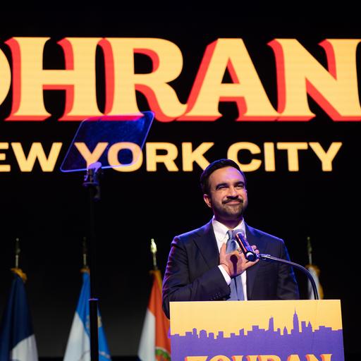 Ein Mann steht auf einer Bühne und fasst sich mit der rechten Hand aufs Herz. Es ist der neu gewählte New Yorker Bürgermeister Zohran Mamdani. Er hat einen Bart, hinter ihm ist ein Plakat mit seinem Vornamen aufgehängt.