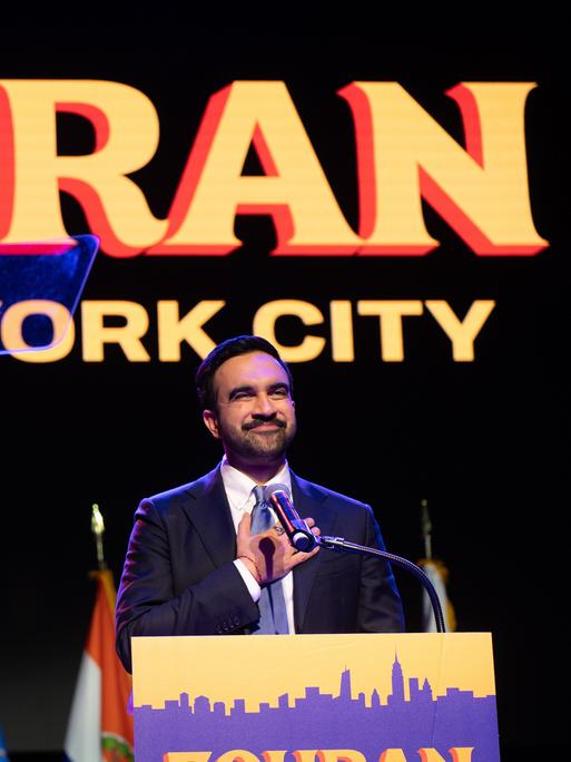 Ein Mann steht auf einer Bühne und fasst sich mit der rechten Hand aufs Herz. Es ist der neu gewählte New Yorker Bürgermeister Zohran Mamdani. Er hat einen Bart, hinter ihm ist ein Plakat mit seinem Vornamen aufgehängt. Ein Mann steht auf einer Bühne und fasst sich mit der rechten Hand aufs Herz. Es ist der neu gewählte New Yorker Bürgermeister Zohran Mamdani. Er hat einen Bart, hinter ihm ist ein Plakat mit seinem Vornamen aufgehängt.