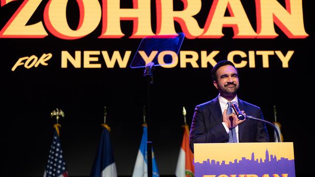 Ein Mann steht auf einer Bühne und fasst sich mit der rechten Hand aufs Herz. Es ist der neu gewählte New Yorker Bürgermeister Zohran Mamdani. Er hat einen Bart, hinter ihm ist ein Plakat mit seinem Vornamen aufgehängt.