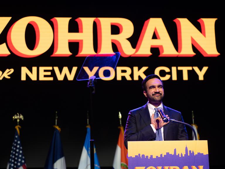 Ein Mann steht auf einer Bühne und fasst sich mit der rechten Hand aufs Herz. Es ist der neu gewählte New Yorker Bürgermeister Zohran Mamdani. Er hat einen Bart, hinter ihm ist ein Plakat mit seinem Vornamen aufgehängt. Ein Mann steht auf einer Bühne und fasst sich mit der rechten Hand aufs Herz. Es ist der neu gewählte New Yorker Bürgermeister Zohran Mamdani. Er hat einen Bart, hinter ihm ist ein Plakat mit seinem Vornamen aufgehängt.