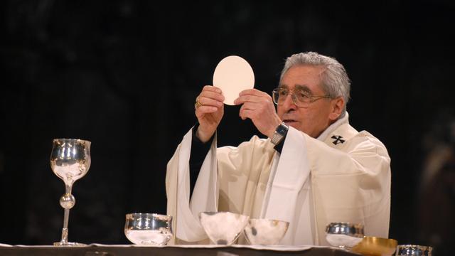 Erzbischof Jean-Marie Lustiger bei einer Eucharistiefeier. Er steht in weißem Gewand am Altar, hält eine Hostie hoch. Neben ihm steht ein Weinbecher und