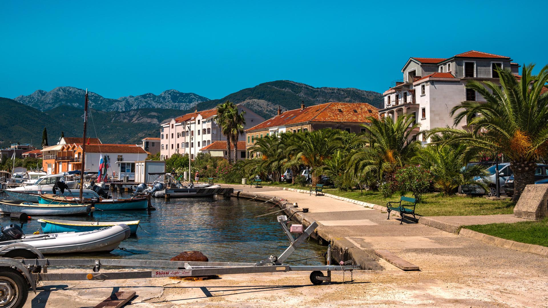 Blick von der Marina auf den Küstenort Tivat in Montenegro. Einige Boote liegen im Wasser, es gibt eine Strandpromenade und Häuser vor blauem Himmel im Hintergrund. 