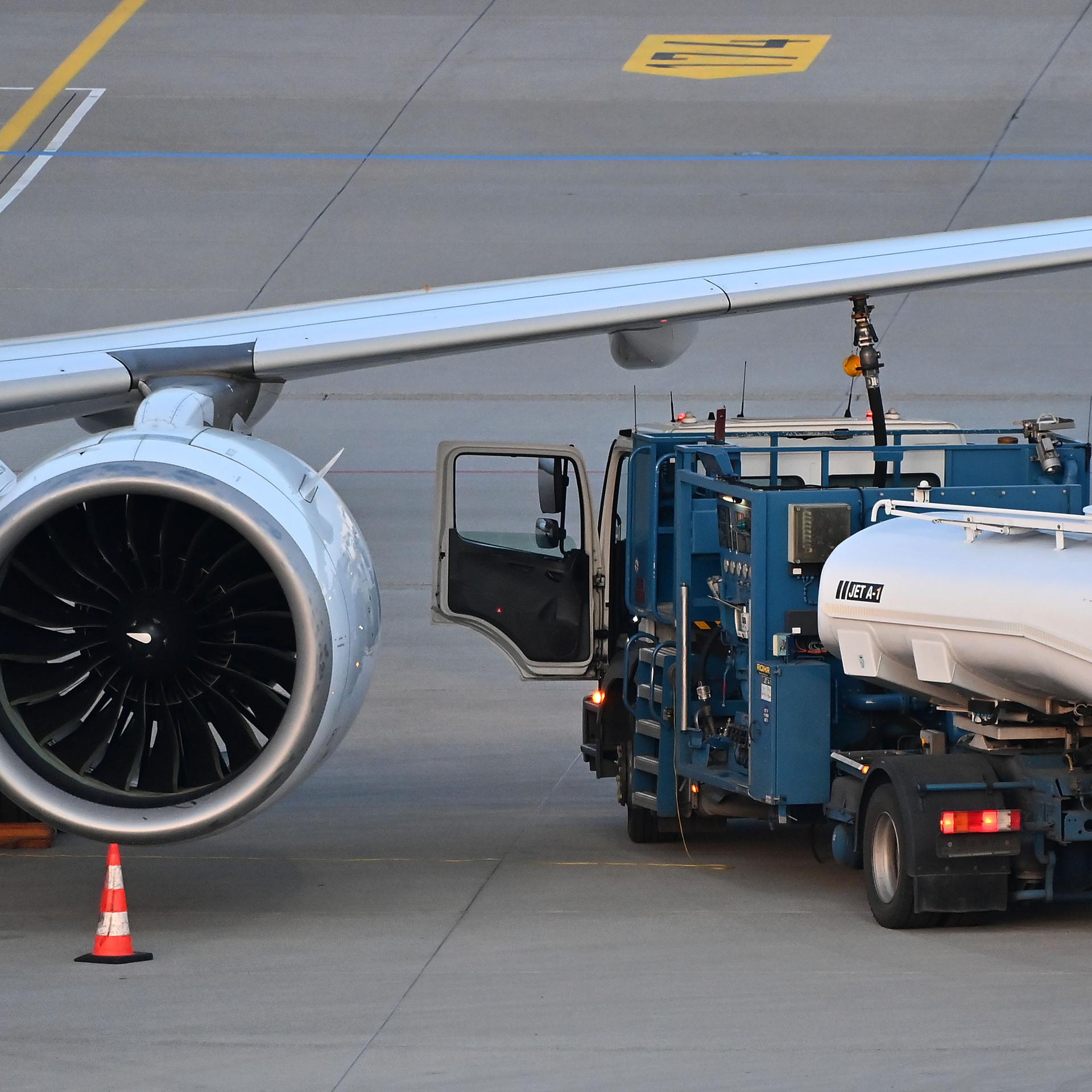 Von dem Flugzeug sieht man nur einen Flügel mit einer großen Turbine. Daneben steht das Tankfahrzeug, aus dem ein Schlauch mit der Tanköffnung im Flügel verbunden ist. 
