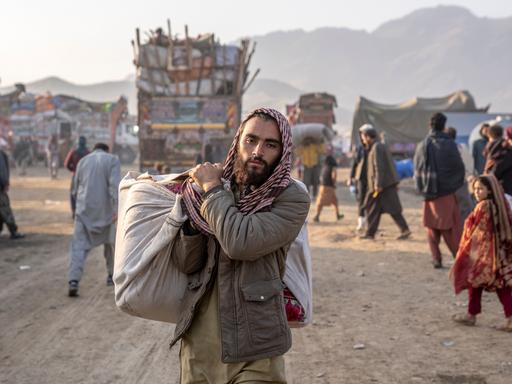 Ein geflüchteter Afghane steht in Pakistan an einem Grenzübergang. Er trägt Habseligkeiten bei sich und blickt dabei in die Kamera.