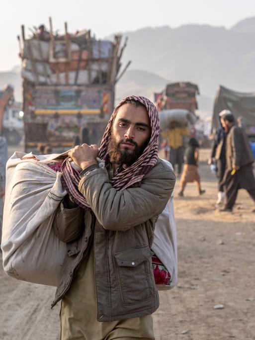 Ein geflüchteter Afghane steht in Pakistan an einem Grenzübergang. Er trägt Habseligkeiten bei sich und blickt dabei in die Kamera.