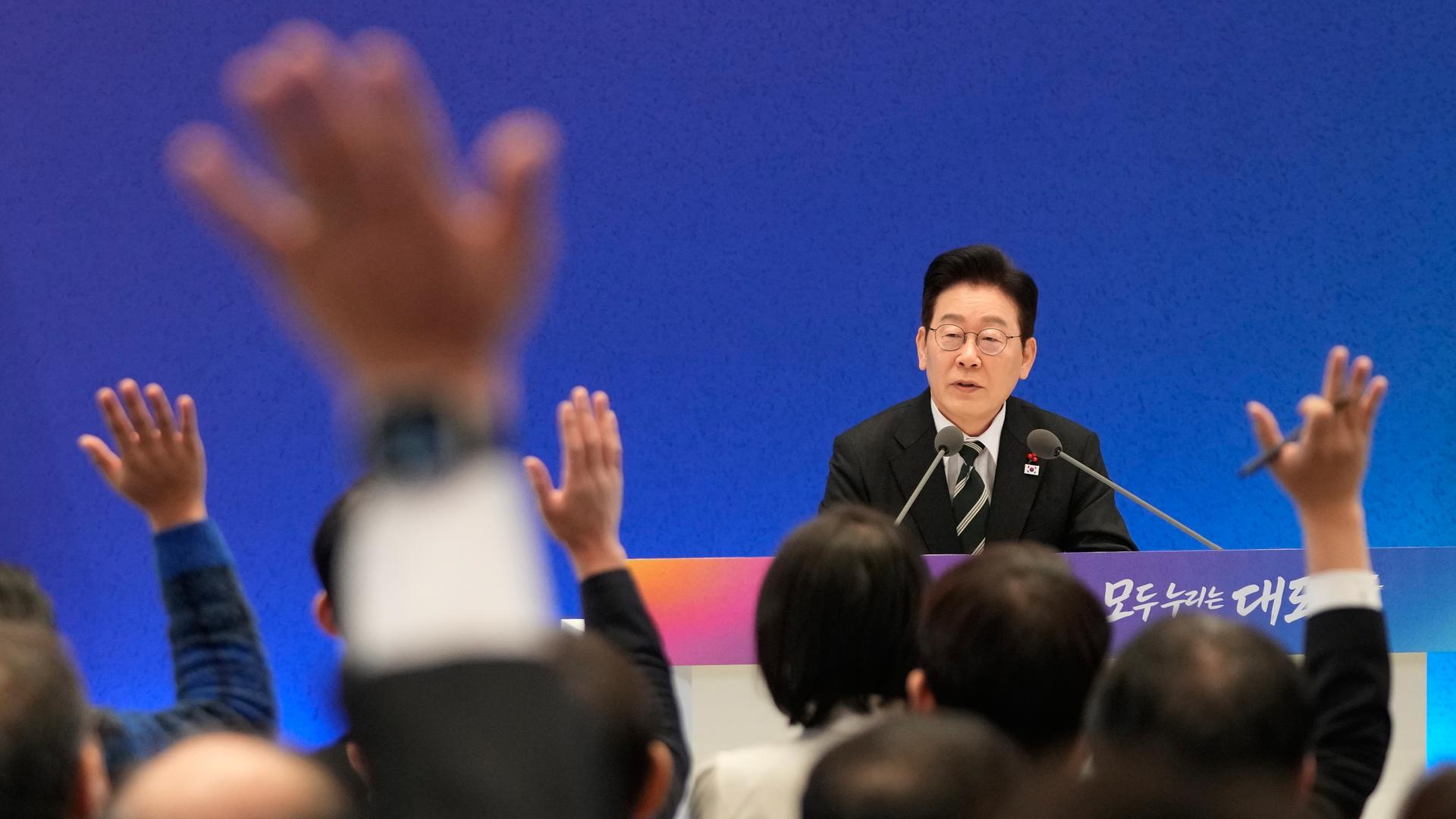 Der südkoreanische Präsident Lee Jae Myung sitzt in einer Pressekonferenz hinter Mikrofonen. Man sieht einige Hände, die in die Höhe gehalten werden.