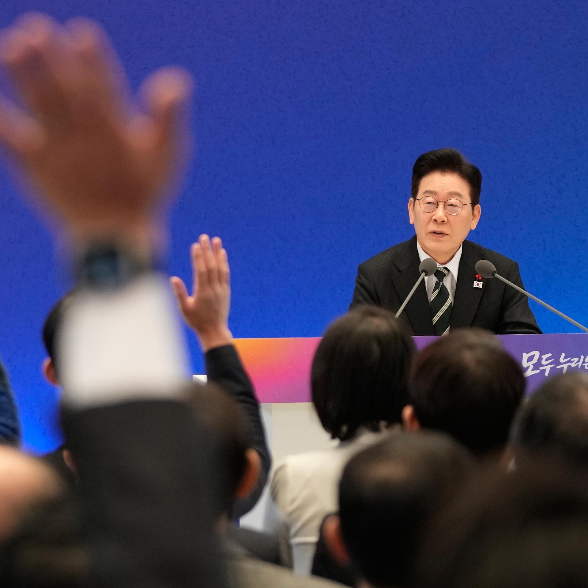 Der südkoreanische Präsident Lee Jae Myung sitzt in einer Pressekonferenz hinter Mikrofonen. Man sieht einige Hände, die in die Höhe gehalten werden.
