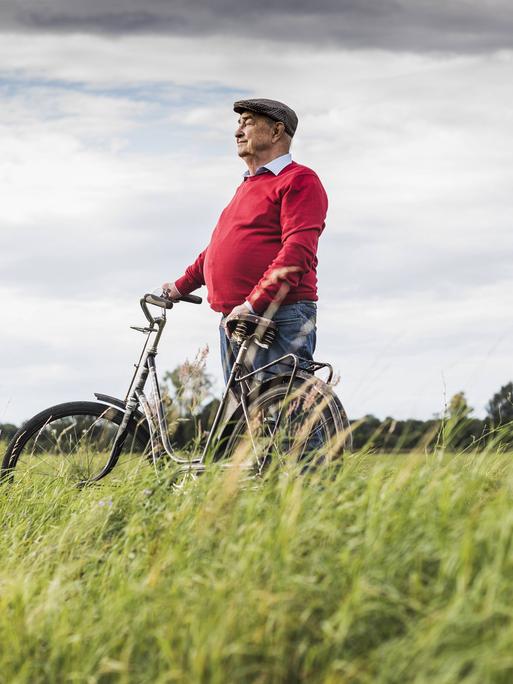 Ein älterer Herr steht mit einem Fahrrad zwischen einem Weizenfeld und Wald. 