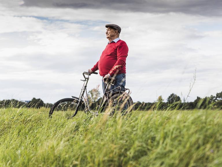 Ein älterer Herr steht mit einem Fahrrad zwischen einem Weizenfeld und Wald. 