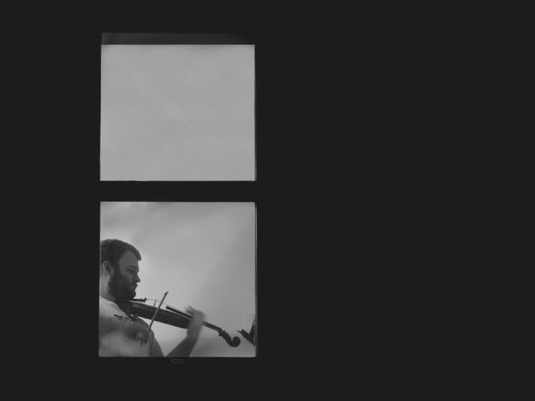 Ein Geiger spielt in einem hell erleuchteten Fenster Geige, das inmitten einer dunklen Fassade liegt.