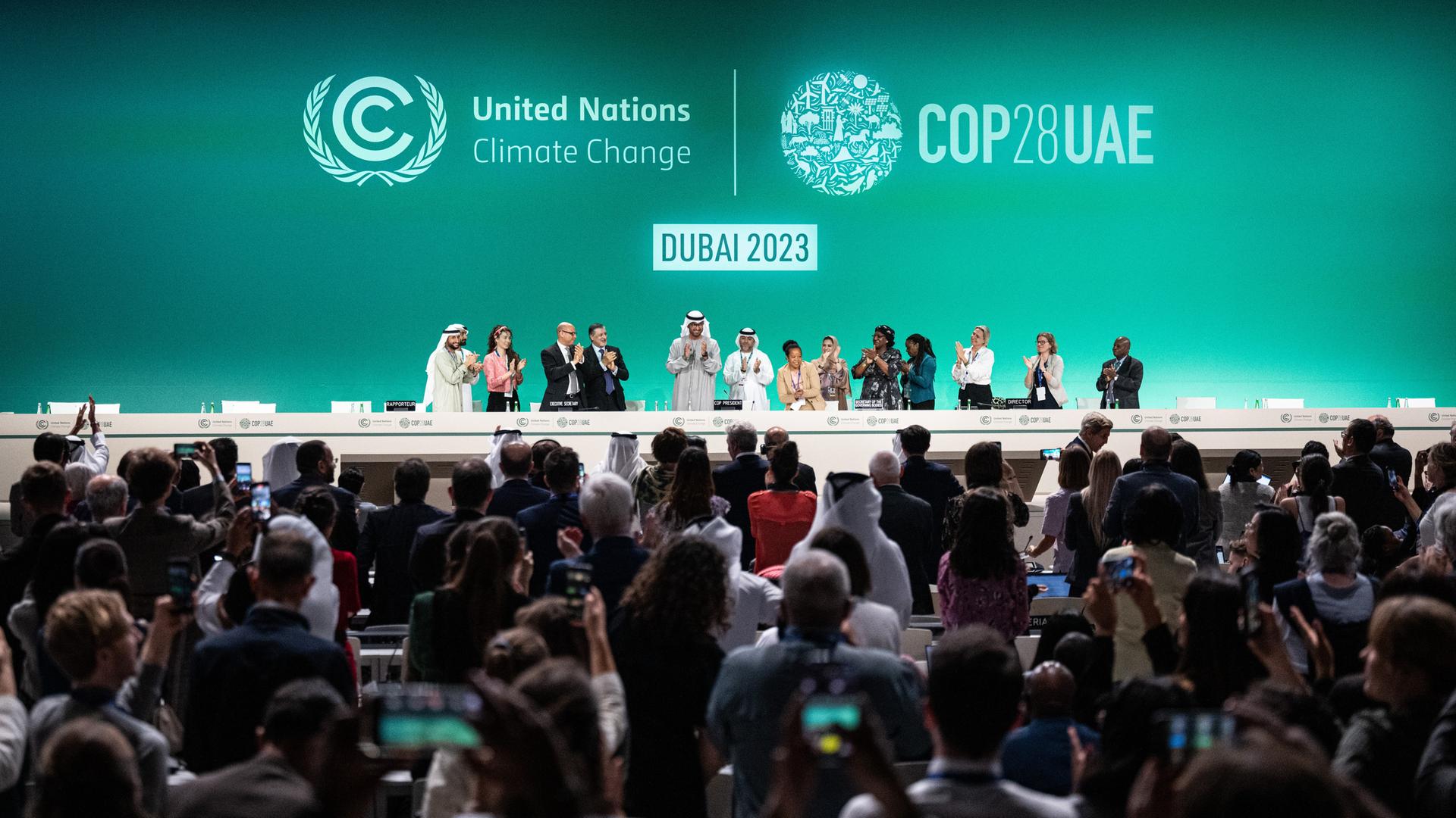 Blick auf das Podium der COP28 - Vertreter unterschiedlicher Länder stehen und applaudieren