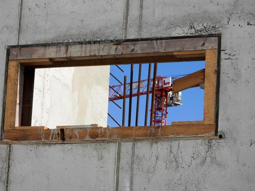 Blick durch eine Fensterlücke im Rohbau eines Wohnhauses mit Krahn im Hintergrund