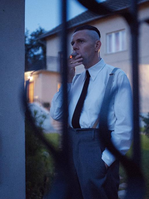 Der Darsteller steht Zigarre rauchend hinter einem Zaun. Er trägt ein weißes Hemd und schwarzen Schlips. Im Hintergrund ist ein Haus zu sehen.