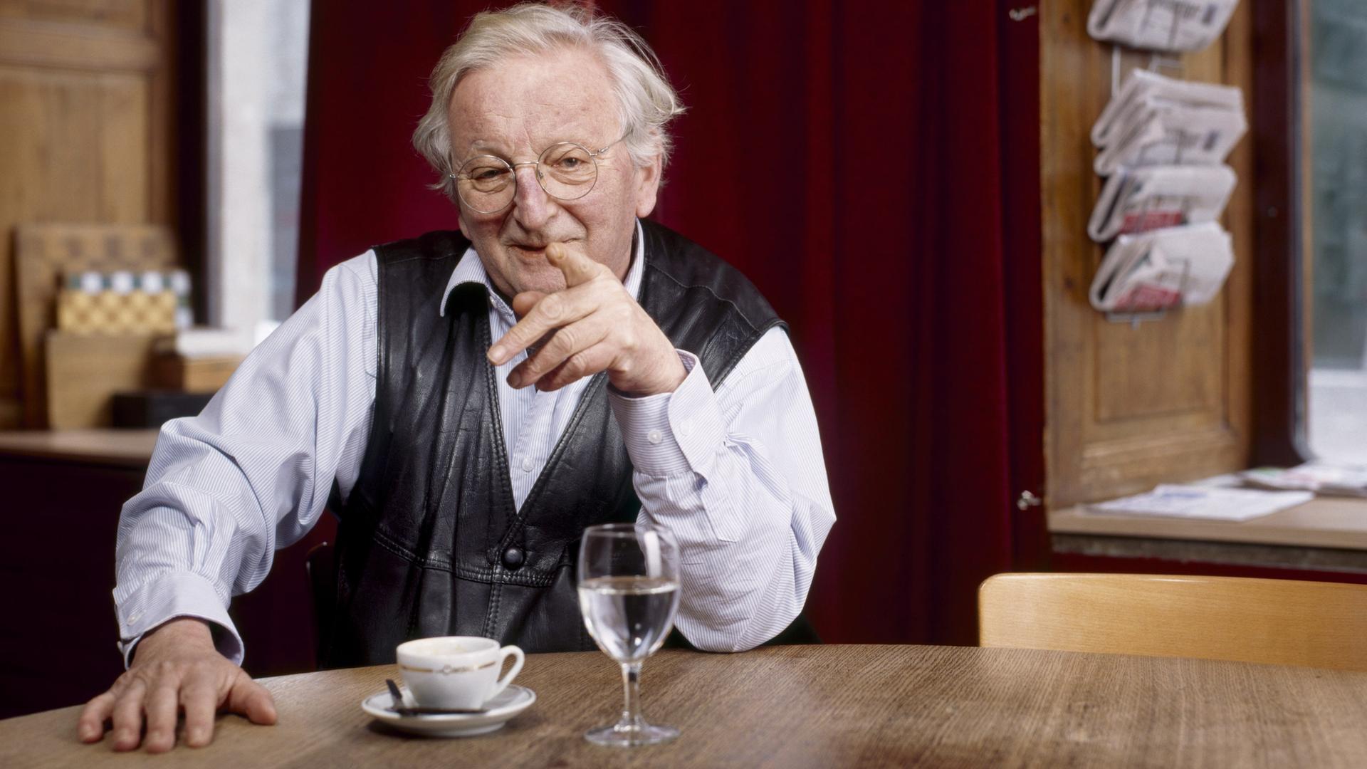 Portrait von Peter Bichsel, Schweizer Schriftsteller. Er sitzt an einem Holztisch unter einer weißen Lampe und hebt den Finger. 