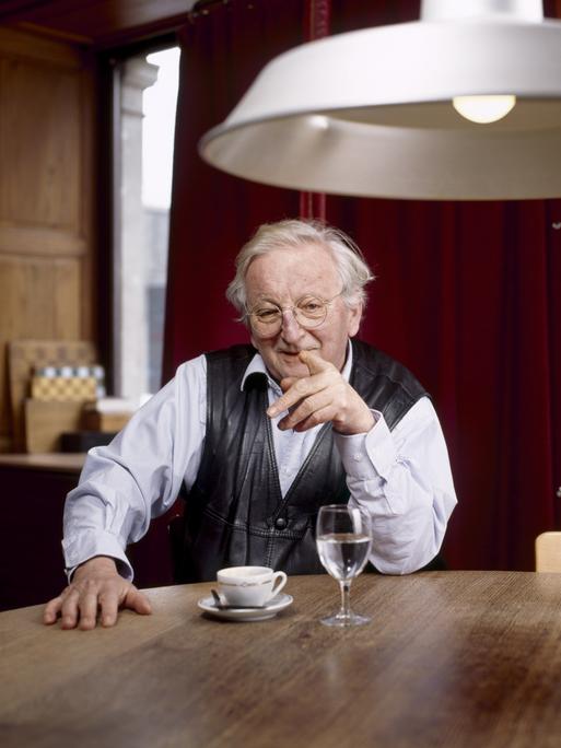 Portrait von Peter Bichsel, Schweizer Schriftsteller. Er sitzt an einem Holztisch unter einer weißen Lampe und hebt den Finger. 