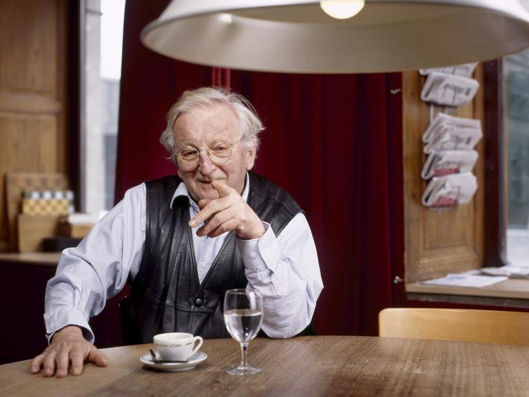 Portrait von Peter Bichsel, Schweizer Schriftsteller. Er sitzt an einem Holztisch unter einer weißen Lampe und hebt den Finger. 