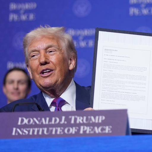 Donald Trump präsentiert eine unterzeichnete Urkunde. Die Platzkarte vor ihm trägt die Aufschrift "Donald J. Trump - Institute of Peace".
