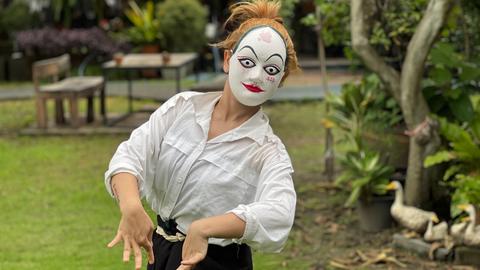 Eine junge Frau bewegt sich - auf dem Gesicht weiße bemalte Papp-Maske - rhythmisch durch einen Garten.
