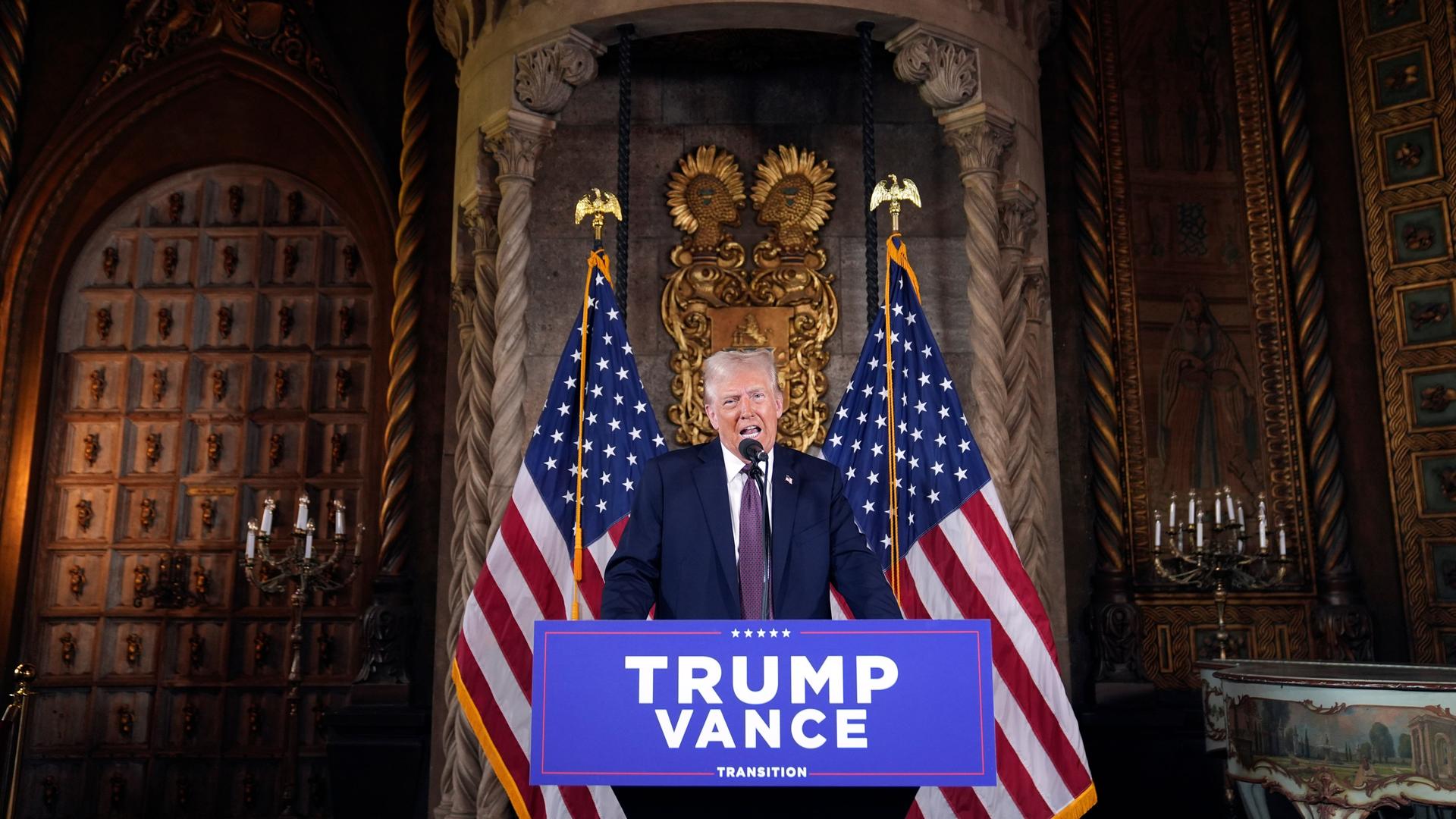 Der künftige US-Präsident Donald Trump am Rednerpult bei einer Pressekonferenz in  Florida. Hinter ihm sind zwei US-Flaggen sowie eine palastähnliche Umgebung zu sehen. Vor ihm hängt ein Schild mit den Namen Trump und Vance.  