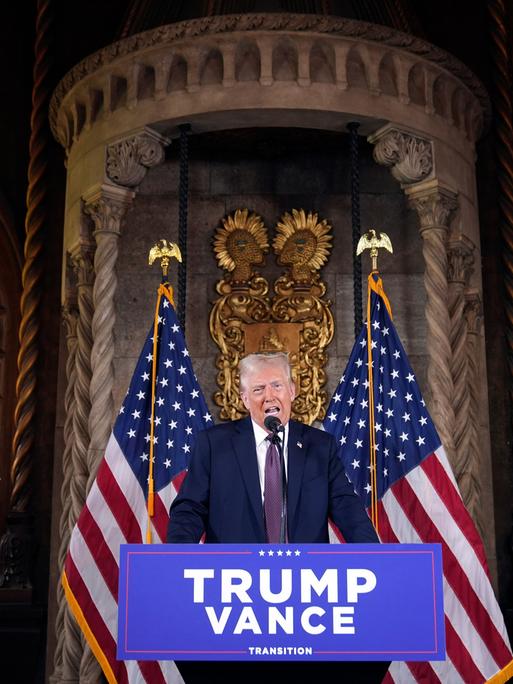 Der künftige US-Präsident Donald Trump am Rednerpult bei einer Pressekonferenz in  Florida. Hinter ihm sind zwei US-Flaggen sowie eine palastähnliche Umgebung zu sehen. Vor ihm hängt ein Schild mit den Namen Trump und Vance.  