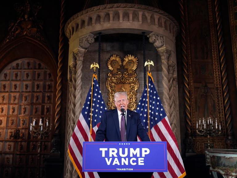 Der künftige US-Präsident Donald Trump am Rednerpult bei einer Pressekonferenz in  Florida. Hinter ihm sind zwei US-Flaggen sowie eine palastähnliche Umgebung zu sehen. Vor ihm hängt ein Schild mit den Namen Trump und Vance.  