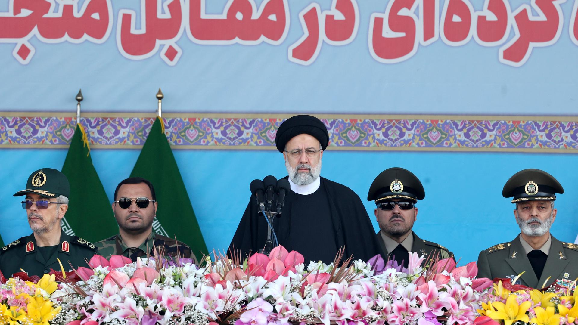 Der iranische Präsident Raisi steht am Militärfeiertag an einem mit Blumen geschmückten Rednerpult. Hinter ihm stehen mehrere Militärs.