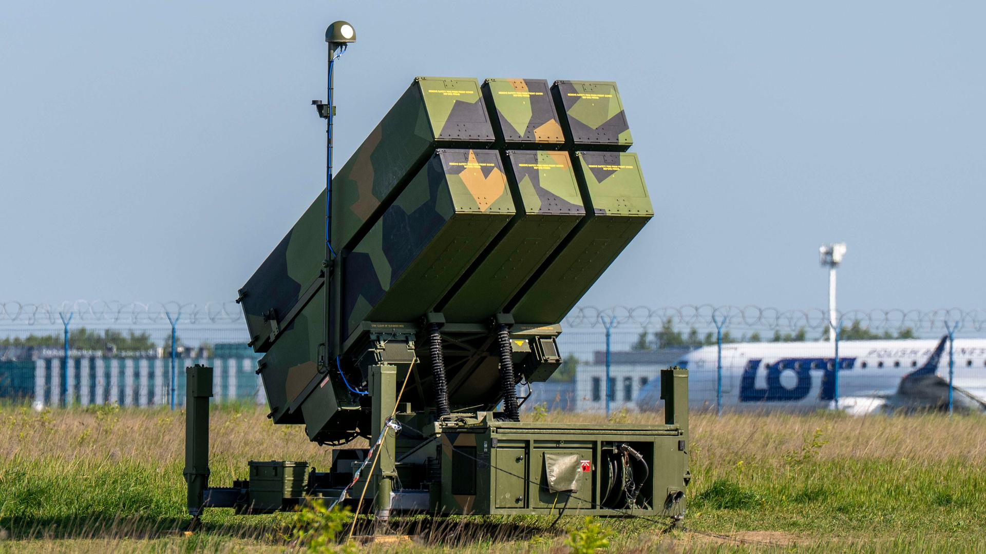Eine in Flecktarn lackierte NASAMS-Einheit steht an einem Rollfeld, im Hintergrund ist ein ziviles Flugzeug der polnischen Fluggesellschaft LOT zu erkennen. Eine in Flecktarn lackierte NASAMS-Einheit steht an einem Rollfeld, im Hintergrund ist ein ziviles Flugzeug der polnischen Fluggesellschaft LOT zu erkennen.