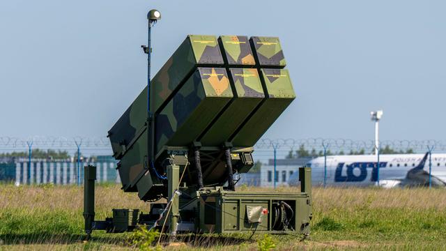 Eine in Flecktarn lackierte NASAMS-Einheit steht an einem Rollfeld, im Hintergrund ist ein ziviles Flugzeug der polnischen Fluggesellschaft LOT zu erkennen.
