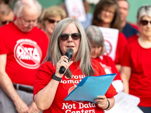 Holly Manke hält im Juli 2026 in Virginia (USA) während einer Kundgebung gegen Rechenzentren eine Rede. Sie trägt ein rotes Tshirt mit der Aufschrift "Grow Tomatos, not Data Centers". Hinter ihr stehen weitere Protestierende.