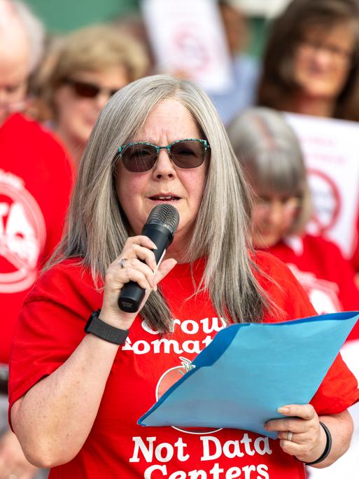 Holly Manke hält im Juli 2026 in Virginia (USA) während einer Kundgebung gegen Rechenzentren eine Rede. Sie trägt ein rotes Tshirt mit der Aufschrift "Grow Tomatos, not Data Centers". Hinter ihr stehen weitere Protestierende.