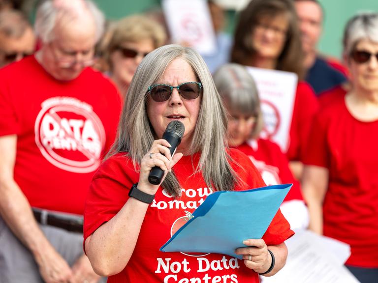 Holly Manke hält im Juli 2026 in Virginia (USA) während einer Kundgebung gegen Rechenzentren eine Rede. Sie trägt ein rotes Tshirt mit der Aufschrift "Grow Tomatos, not Data Centers". Hinter ihr stehen weitere Protestierende.