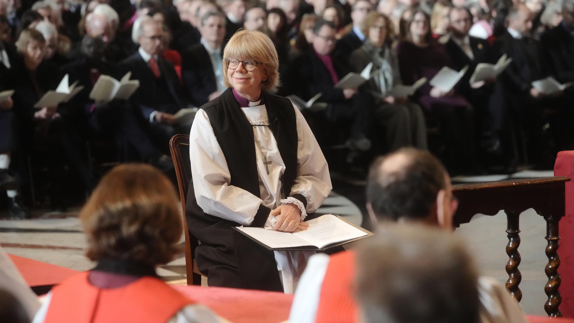 Mullally sitzt lächelnd in einem Stuhl auf dem Chor einer Kirche und blickt zur Seite. Davor unscharf weitere Sitzende, im Hintergrund viele Kirchgänger auf den Bänken. 