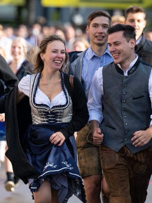 Die ersten Wiesnbesucher rennen nach dem Einlass zu den Festzelten. Im Vordergrund ist eine lachende, junge Frau im Dirndl zu sehen, rechts neben ihr ein junger Mann in Lederhose und Weste über weißem Hemd. Auch die Menschen im Hintergrund strahlen voller Vorfreude auf den Wiesnbesuch. Links im Bild richtet ein junger Mann in Lederhose und schwarzem Hoodie ein Handy auf sich.