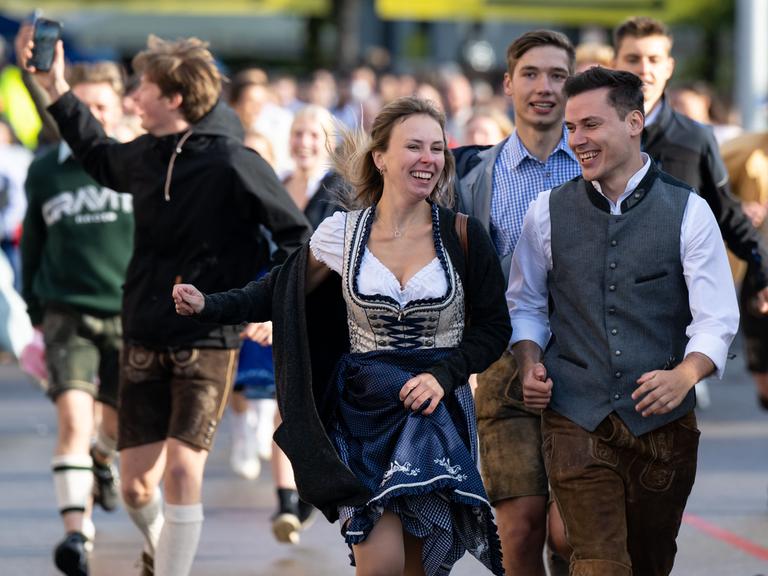 Die ersten Wiesnbesucher rennen nach dem Einlass zu den Festzelten. Im Vordergrund ist eine lachende, junge Frau im Dirndl zu sehen, rechts neben ihr ein junger Mann in Lederhose und Weste über weißem Hemd. Auch die Menschen im Hintergrund strahlen voller Vorfreude auf den Wiesnbesuch. Links im Bild richtet ein junger Mann in Lederhose und schwarzem Hoodie ein Handy auf sich.