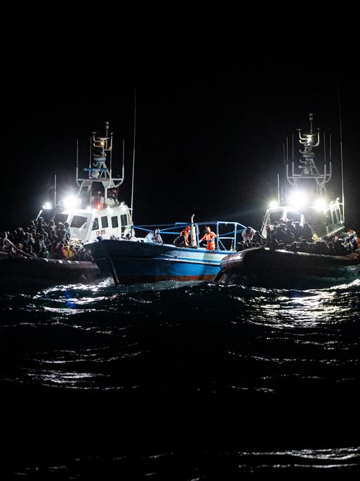 Nachts auf dem Meer: Zwei Boote mit Aufbauten und Scheinwerfern, auf denen sich Menschen drängen, flankieren ein niedriges, offenes Boot.