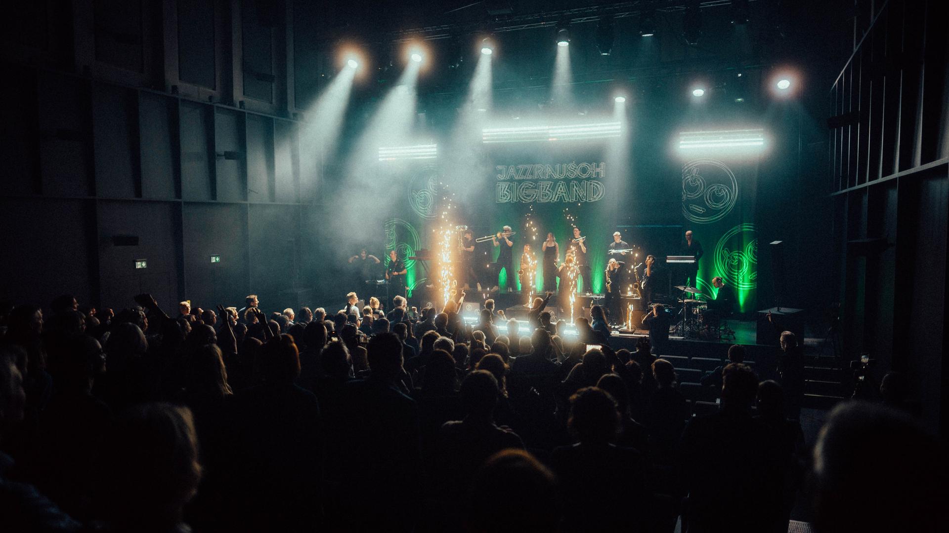 Ein dunkler Konzertsaal, auf der hell beleuchteten Bühne spielt die Jazzrausch Bigband zwischen Lichtfontänen. Ein dunkler Konzertsaal, auf der hell beleuchteten Bühne spielt die Jazzrausch Bigband zwischen Lichtfontänen.
