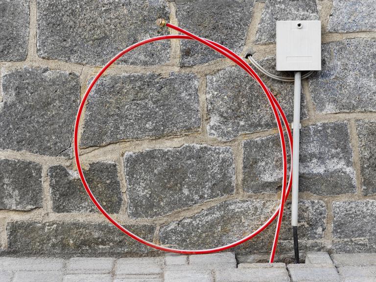 Das Foto zeigt einenTelekom- Hausanschluss mit Glasfaserkabel: Ein gerolltes Kabel lehnt an einer Mauer, daneben ein Verteilerkasten.