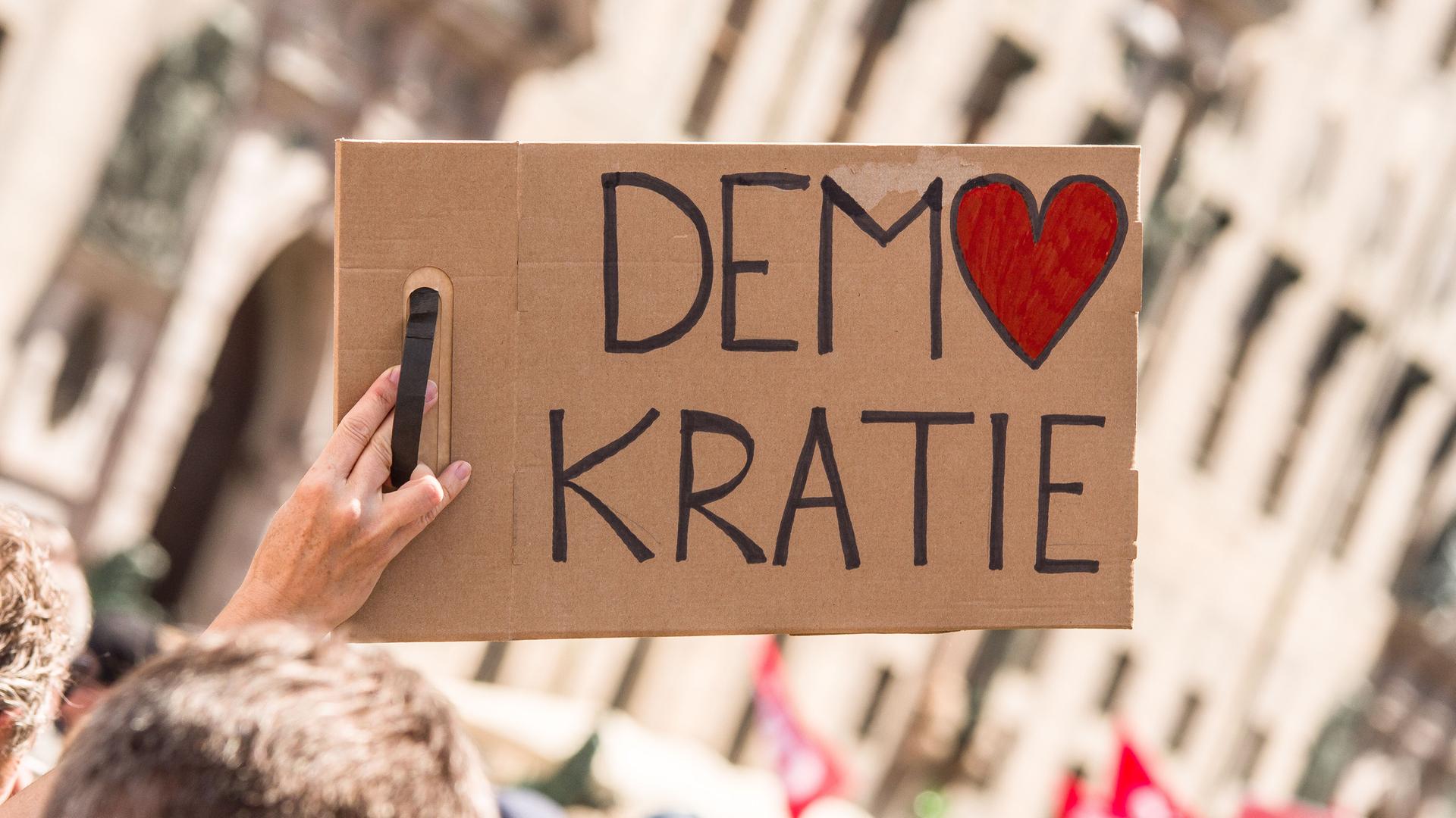 Bei einer Kundgebung wird ein Schild mit der Aufschrift "Demokratie" in die Höhe gestreckt. Dabei ist das O in Demokratie in Form eines Herzes.