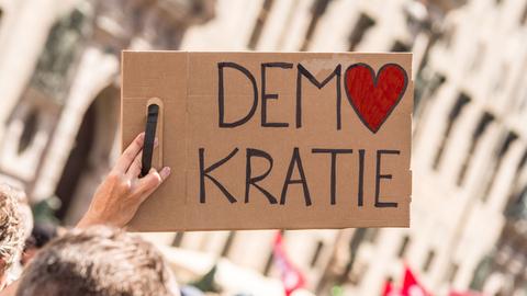 Bei einer Kundgebung wird ein Schild mit der Aufschrift "Demokratie" in die Höhe gestreckt. Dabei ist das O in Demokratie in Form eines Herzes. Bei einer Kundgebung wird ein Schild mit der Aufschrift "Demokratie" in die Höhe gestreckt. Dabei ist das O in Demokratie in Form eines Herzes.