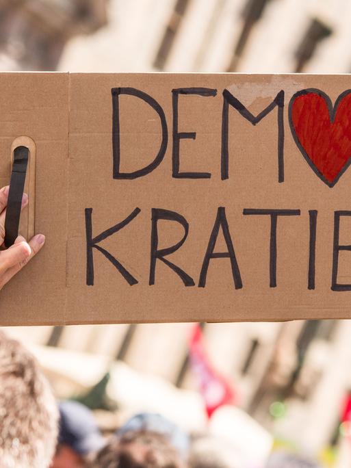Bei einer Kundgebung wird ein Schild mit der Aufschrift "Demokratie" in die Höhe gestreckt. Dabei ist das O in Demokratie in Form eines Herzes.
