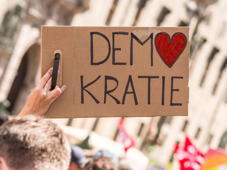 Bei einer Kundgebung wird ein Schild mit der Aufschrift "Demokratie" in die Höhe gestreckt. Dabei ist das O in Demokratie in Form eines Herzes.