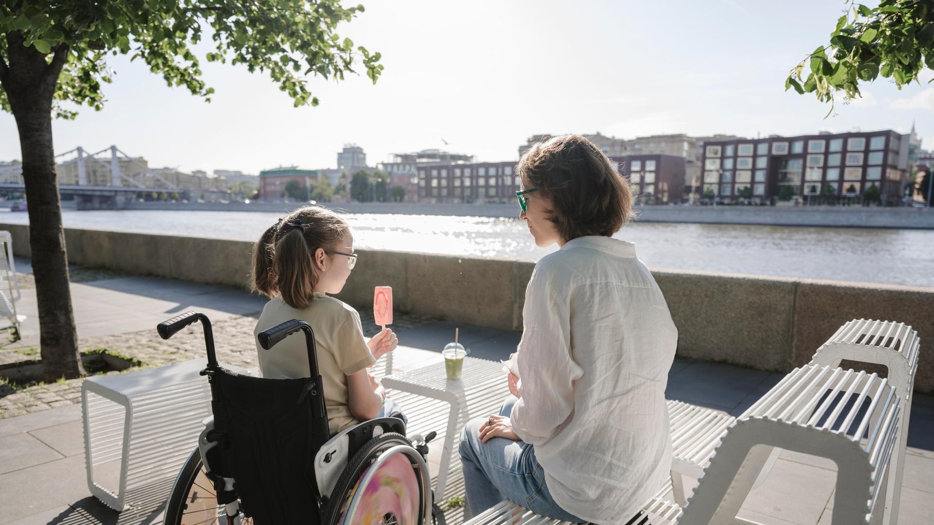 Ein Mädchen im Rollstuhl isst ein Eis, daneben sitzt eine Frau auf einer Bank Ein Mädchen im Rollstuhl isst ein Eis, daneben sitzt eine Frau auf einer Bank