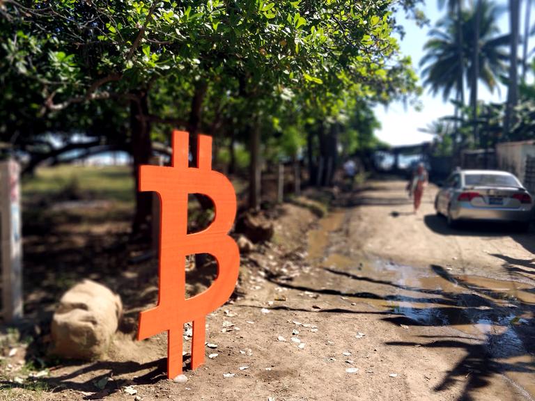 Ein mannshohes Bitcoin-Zeichen steht an einer Straße, die zum Meer führt. 