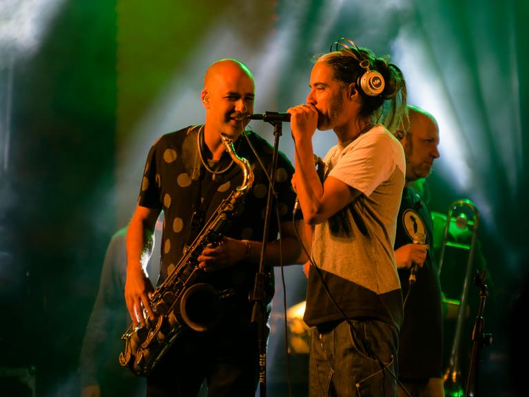 Der Sänger und der Saxophonist der Band stehen einander zugewandt auf der Bühne.